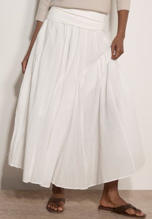 Kobieta nosząca zwiewną białą midi spódnicę i brązowe sandały wsuwane, trzymająca materiał spódnicy po jednej stronie.