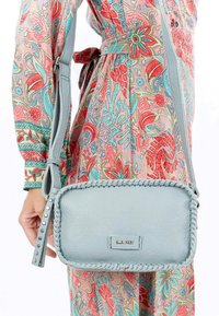 Borsa a tracolla in pelle azzurro chiaro con bordi intrecciati, accessori in argento e una tracolla con motivo, tenuta contro un vestito a stampa floreale.