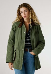 Giacca verde oliva con colletto in suede marrone, bottoni a pressione e tasche, indossata sopra una maglietta a righe blu e bianche e jeans blu.