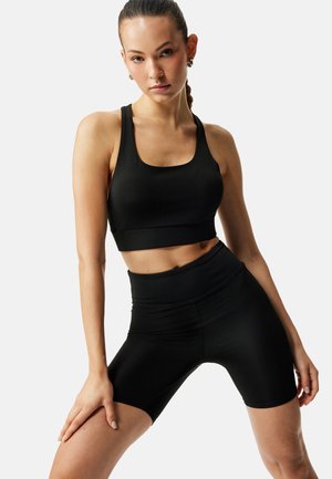 Frau posiert in schwarzem Sport-BH und hoch taillierten Biker-Shorts vor einem schlichten hellen Hintergrund.