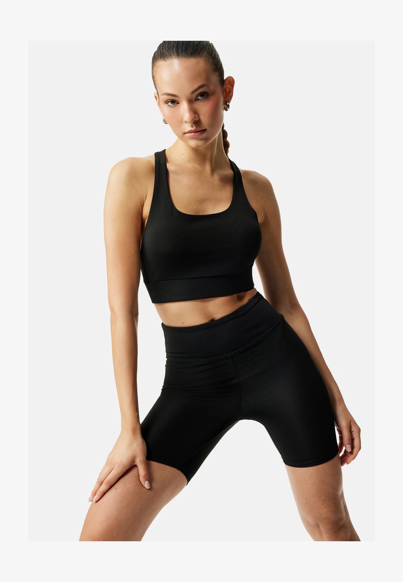 Vrouw poseert in een zwarte sportbeha en hoge bikershorts tegen een effen lichte achtergrond.