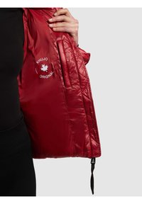 Käsi pitelee avattua punaista tikattua takkia, joka paljastaa sisätaskun ja valkoisen "KHUJO ORIGINAL" -tekstin vaahteranlehtilogon kanssa vuorin sisäpuolella.