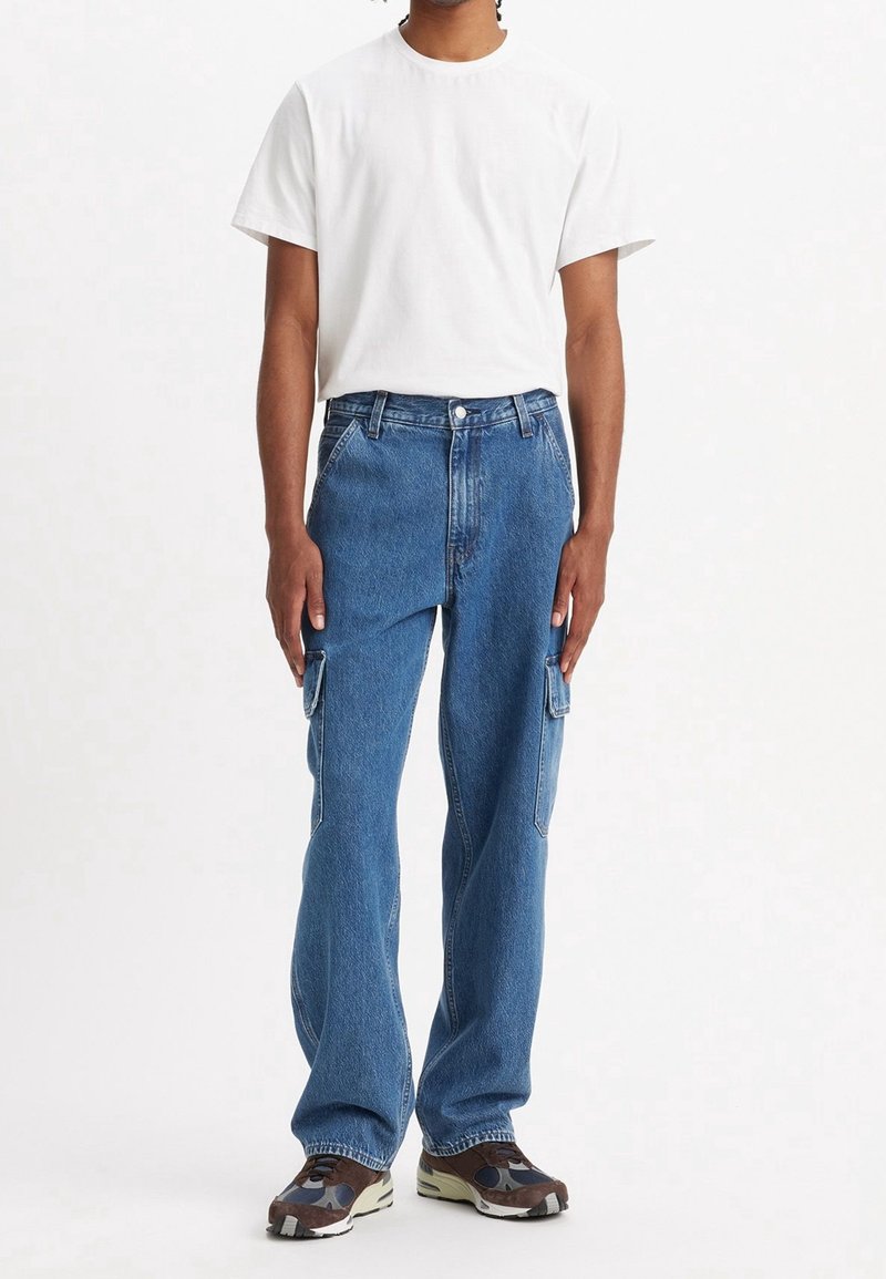 Personne portant une chemise blanche unie à manches courtes, un jean cargo en denim bleu, et des baskets marron et noires, debout devant un fond blanc.