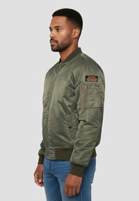 Olijfgroene bomberjas met een ritssluiting aan de voorkant, geribbelde manchetten en zoom; heeft een patch op de mouw en zijzakken. Gladde satijnen textuur.