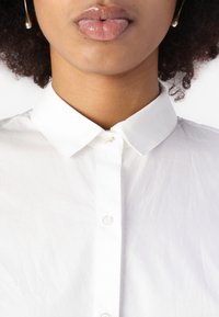 White button-up shirt with a classic collar, featuring a row of buttons and a subtle gold accent at the neckline. Smooth, lightweight fabric.