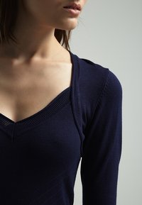 Haut en maille bleu marine avec un col en V profond, bordure côtelée et manches longues. Texture lisse avec des détails de motifs horizontaux subtils.