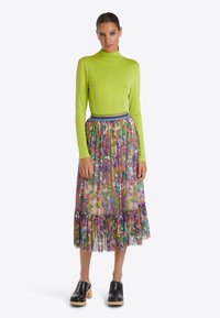 Langarm, tailliertes, leuchtend limettengrünes Oberteil, kombiniert mit einem mehrfarbigen, blumigen, plissierten Rock mit Rüschensaum. Schwarze Clogs runden den Look ab.