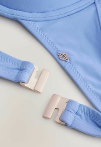 Šviesiai mėlyna bikini viršutinė dalis su lygiu audiniu, turinti rožinio aukso užsegimą ir dekoratyvinį siuvinėjimą. Mažas logotipo detalė prie siūlės.