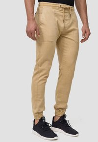 Beige joggerbukser med elastisk talje, snore og sidelommer. Tapered fit med ribbede manchetter ved anklerne, lavet af blødt stof.