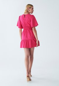 Rosa kurzes Kleid mit glockenförmigen Ärmeln, gerafftem Taillenbund und Rüschensaum. Die Rückseite hat eine Schlüssellochöffnung. Kombiniert mit nudefarbenen High Heels.