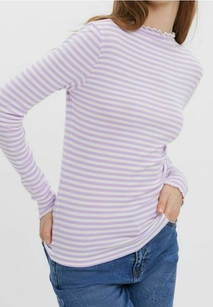 Frau trägt ein langärmeliges, lavendelweiß gestreiftes Oberteil mit Rüschenkanten, kombiniert mit blauen Jeans, mit Händen in den Taschen.