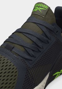 Σκούρο πράσινο και μαύρο αθλητικό παπούτσι Reebok με υφή υφάσματος, μαύρα κορδόνια και νέον πράσινο λογότυπο στη γλώσσα και στη σόλα.