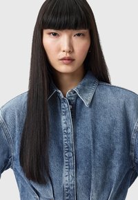 Camicia di denim azzurro chiaro con colletto classico, chiusura a bottoni e design dritto. Tessuto testurizzato con dettagli di cucitura visibili.