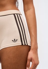 Κοντινό πλάνο ενός ατόμου που φοράει μπεζ σορτς με τρεις μαύρες κάθετες ρίγες και μαύρο λογότυπο τριφυλλιού στη πλευρά, σε απλό φόντο.