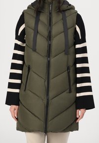 Kobieta ubrana w oliwkowo-zieloną bezrękawnikową pikowaną kamizelkę z zamkiem z przodu, na czarno-białej, paskowanej, długorękawowej bluzce.