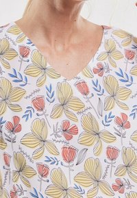Floral-kuvioinen V-pääkalu, jossa on keltaisia ja punaisia kukkia valkoisella taustalla, sinisiä lehtiä ja hienoja mustia ääriviivoja.