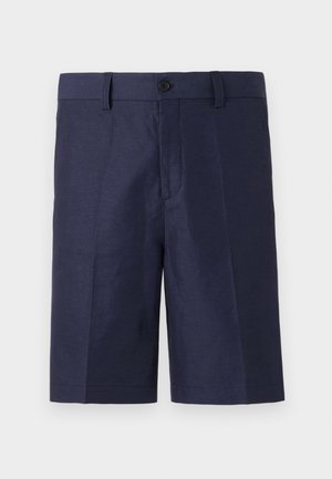 Musta ribbineulottu poolopaita, jossa kontrastivärinen kaulus, tummansiniset shortsit, mustat sukat ja mustat loafers-kengät. Pitää kädessään pientä ruskeaa nahkalaukkua.