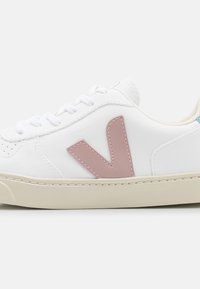 Vit sneaker med en slät yta, prydd med en ljusrosa gummilogotyp i form av ett "V", vita snören och en krämfärgad sula.