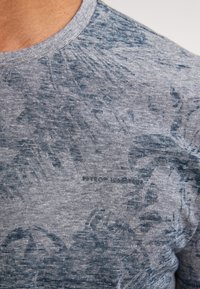 Šedobláškový tričko s kulatým výstřihem vyrobené z texturované tkaniny. Obsahuje jemný vzor listů a malý textový detail u lemu.