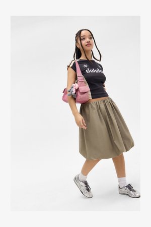 Jonge vrouw met gevlochten haar, gekleed in een zwart graphic T-shirt, beige rok, zilveren sneakers en met een roze schoudertas met sleutelhangeraccessoires.