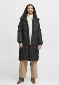 Sort puffet jakke med quiltet design, høj krave, trykknapper og sidelommer. Båret over en cremefarvet turtleneck og beige bukser.