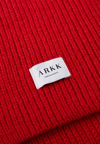 Vörös, kötött anyag függőleges bordázott textúrával, és egy fehér címke, amelyen fekete betűkkel van írva: "ARKK COPENHAGEN".