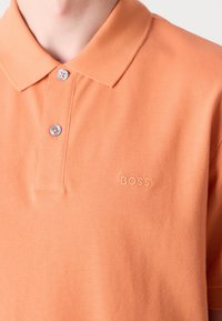 Gros plan d'une personne portant un polo orange à deux boutons avec "BOSS" brodé sur la poitrine.