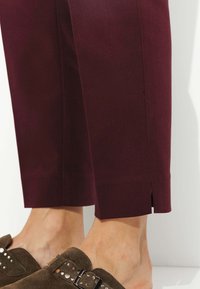 Pantalons bordeaux à coupe ajustée, avec une légère fente à l'ourlet. Associés à des chaussures marron avec des clous décoratifs.
