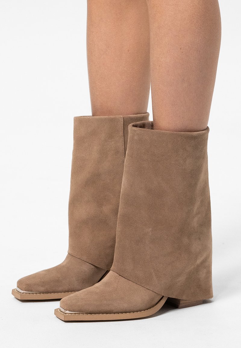 Hohe, taupefarbene Wildlederstiefeletten mit umgeschlagenem Schaft, spitzer Zehenform und flachem Boden. Die Textur ist weich mit einer glatten Oberfläche und minimalen Nähten.