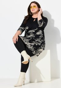 Musta tunikamekko, jossa on paisley- ja kukkakuviota, avaimenreikäneckline, yhdistettynä mustiin legginsseihin ja valkoisiin korkokenkiin nilkoissa.