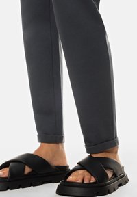 Zwarte sliders met een dikke zool, voorzien van kruislings over elkaar lopende banden. Grijze broek met omslag van zacht materiaal, iets taps toelopend bij de enkels.