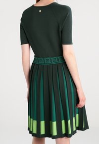 Robe vert foncé à manches courtes, jupe plissée, ourlet contrastant vert clair et taille texturée avec des motifs géométriques.