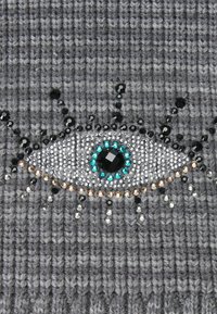 Un tissu tricoté gris présente un motif œil composé de cristaux variés, dont du turquoise et du noir, avec un fond texturé et des accents scintillants.