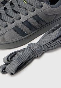 Αθλητικό παπούτσι από γκρι σουέντ με σόλα από σκούρο γκρι καουτσούκ και σχέδιο με τρεις ρίγες. Περιλαμβάνει γκρι κορδόνια με υφή που ταιριάζουν.