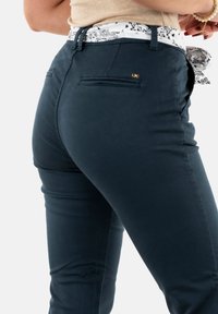 Donkerblauwe broek met een getailleerd ontwerp, voorzien van achterzakken en een contrasterende, geborduurde tailleband. Subtiele logo-details zijn aanwezig.
