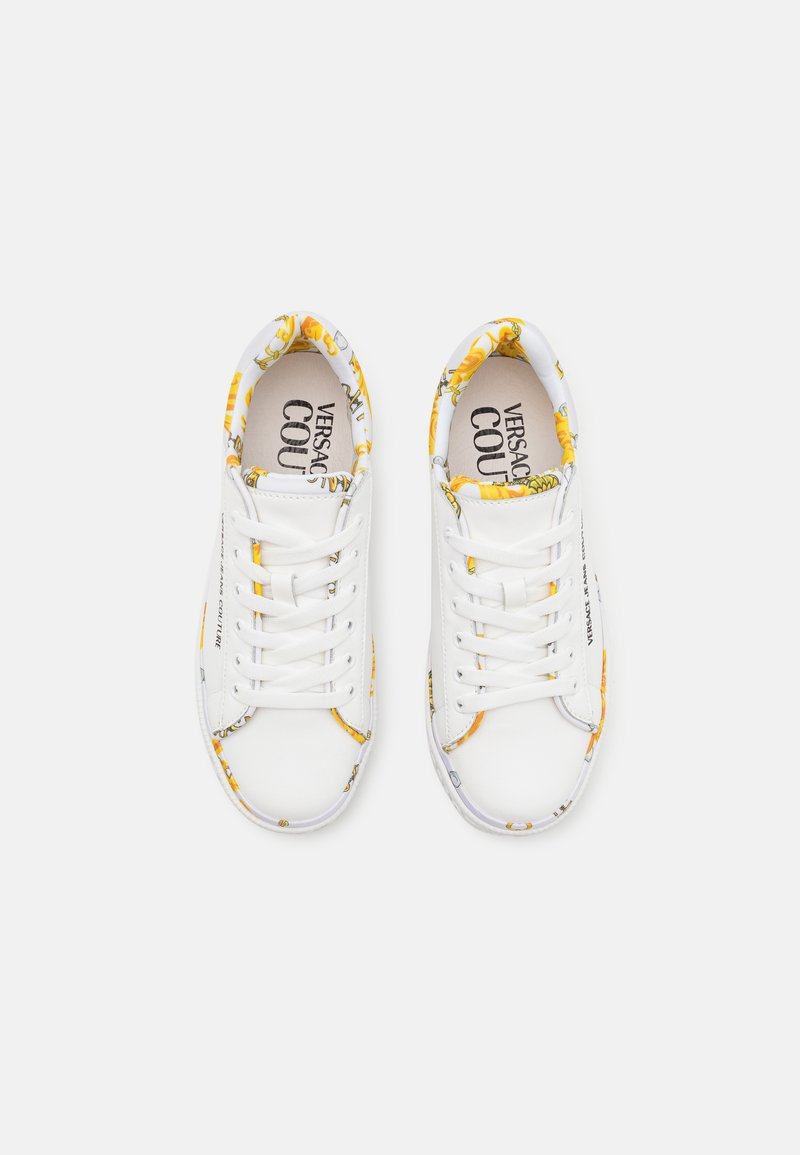 Zapatillas blancas con acentos florales amarillos, puntera redonda, cordones planos, logotipo en la plantilla y ribete decorado a lo largo del cuello y el lateral.