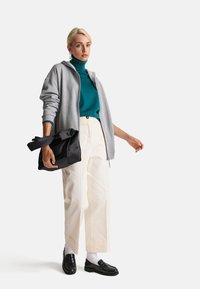 Grijze rits hoodie over een teal coltrui, gecombineerd met crèmekleurige wijdlopende broeken en zwarte loafers, terwijl er een zwarte tote tas wordt vastgehouden.