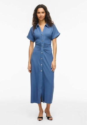 Kobieta o kręconych włosach, ubrana w niebieską sukienkę midi na guziki z krótkim rękawem, marszczoną w talii i czarne sandały na obcasie z odkrytymi palcami, stojąca na białym tle.