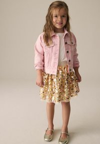 Veste en denim rose sur un T-shirt blanc, associée à une jupe en sequins dorés. Les chaussures sont métalliques avec une bride. Design simple et jeune.