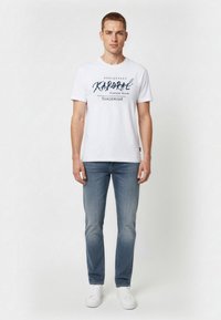 Biała bawełniana koszulka z ciemnoniebieskim nadrukiem tekstu "Kaporal" i logo, zestawiona z jasnoniebieskimi dżinsami i białymi trampkami.