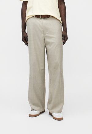 Άνδρας με μπεζ παντελόνι φαρδιάς γραμμής, καφέ ζώνη, εκρού πουκάμισο και λευκά παπούτσια με καφέ σόλες, στέκεται μπροστά σε απλό φόντο.