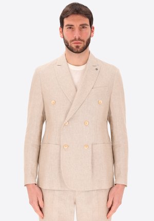 Uomo che indossa un completo in lino beige doppiopetto con sei bottoni, in piedi davanti a uno sfondo chiaro e uniforme, rivolto in avanti.