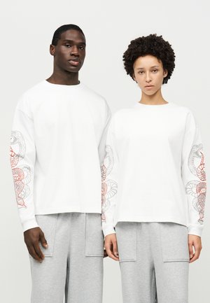 Deux mannequins posent avec des hauts blancs assortis à manches longues, ornés de motifs de serpents sur les manches, et des pantalons de survêtement gris clair.