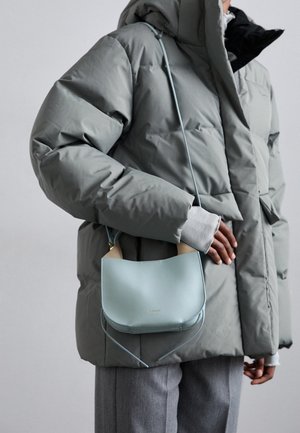 Persoon in een grijze donzen jas en grijze broek draagt een kleine lichtblauwe crossbody tas met beige handvatten.