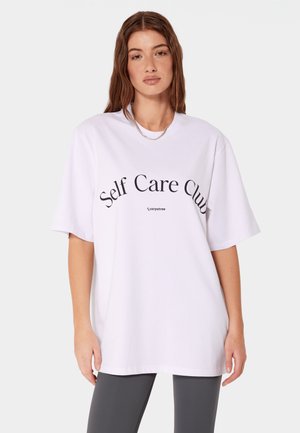 Kobieta z długimi brązowymi włosami, ubrana w luźny biały T-shirt z napisem „Self Care Club” i ciemne legginsy, stojąca na jednolitym jasnym tle.