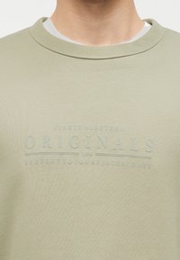 Φωτεινό πράσινο φούτερ με λαιμόκοψη crewneck με ανάγλυφο κείμενο "Ninety Nineteen Originals 1990 Brought to you by Jack & Jones" στο στήθος.