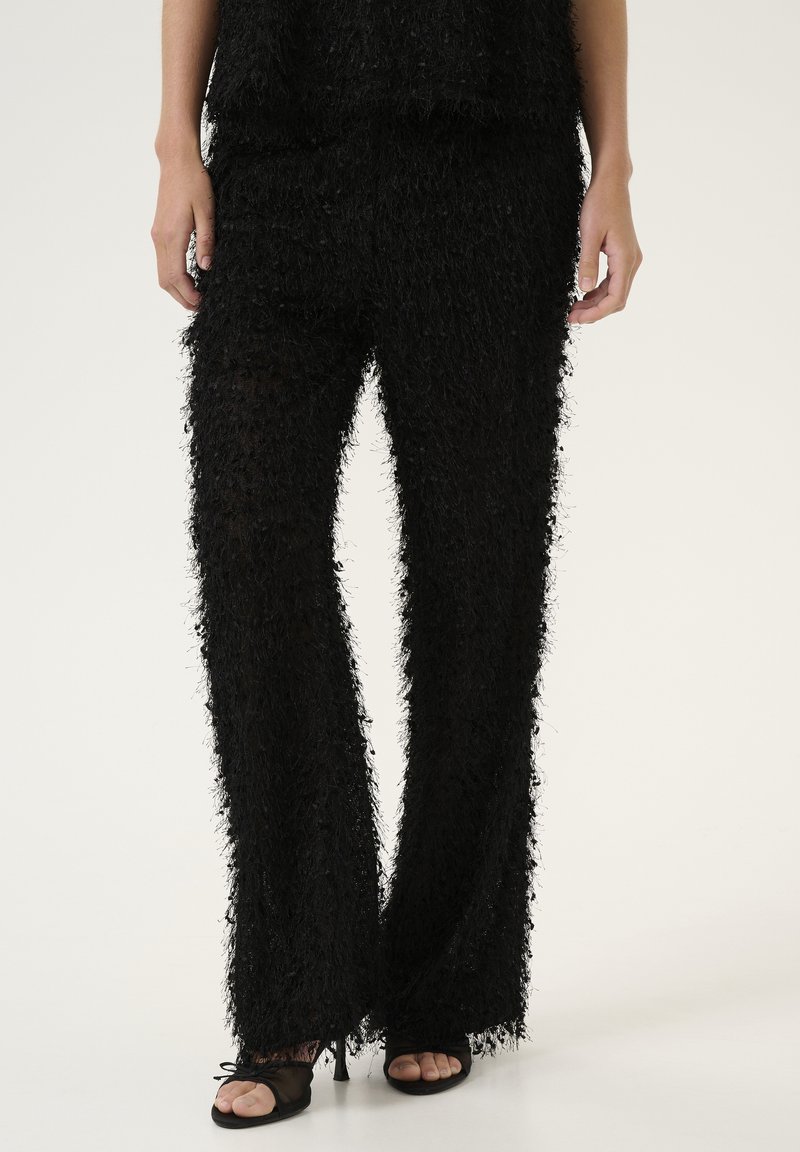 Pantalons noirs ajustés en tissu texturé et duveteux ; forme évasée ; bords effilochés ; associés à des chaussures à bout ouvert noires.