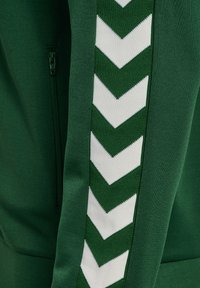 Vihreä kangashuppu, jossa pystysuoria valkoisia kalastusrivistöjä ja vihreä vetoketju vasemmalla puolella.