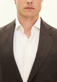 Blazer marrone su misura sopra una camicia bianca con bottoni, dotata di un colletto standard e bottoni visibili, con un sottile dettaglio sulla tasca.