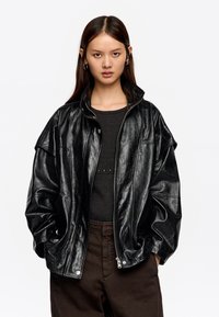 Joven con cabello largo y oscuro, vistiendo una chaqueta de piel negra brillante y oversize, una camiseta negra acanalada y pantalones marrón oscuro, con las manos en los bolsillos.